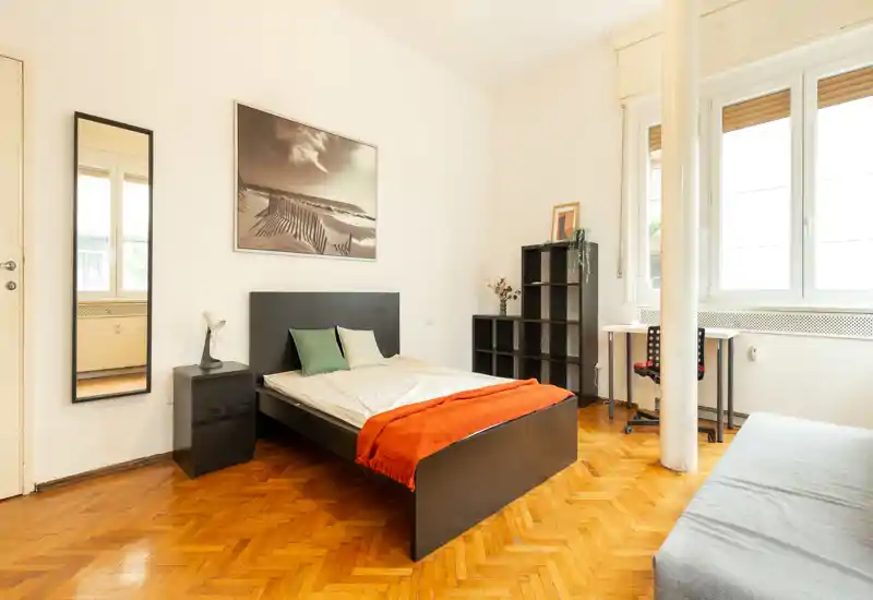 Bright, simply styled bedroom with a double bed, bedside table, full-length mirror and workspace by the window — warm wooden floors and an orange throw add character.