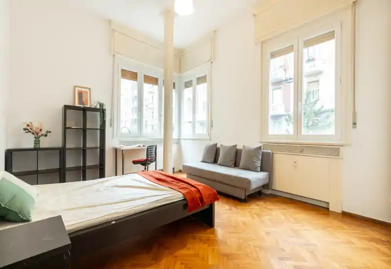 Bright, well-staged bedroom with a double bed, cosy throw and cushions, a small sofa and large windows that let in generous natural light.