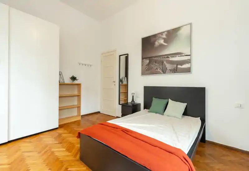 Spacious, bright bedroom with a double bed, parquet flooring and minimalist furnishings. Neutral walls, framed seascape artwork and a warm orange throw create a welcoming atmosphere.