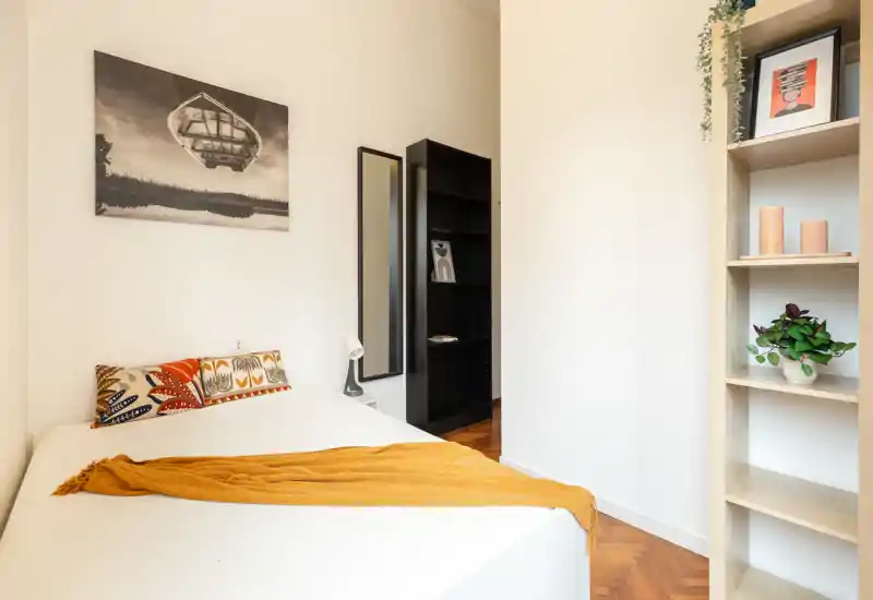 Bright, minimalist bedroom with a comfortable double bed, warm throw and patterned cushions; simple shelving and a mirror add practical storage and depth.