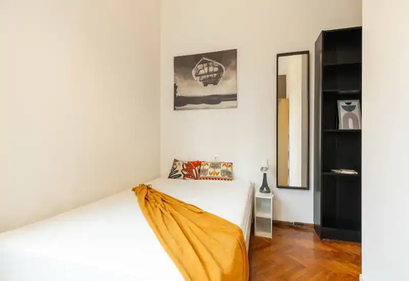 Bright, cozy bedroom with a neatly made double bed, decorative pillows and a warm mustard throw. Simple, modern decor with a full-length mirror and shelving, ideal for a comfortable short-term stay.