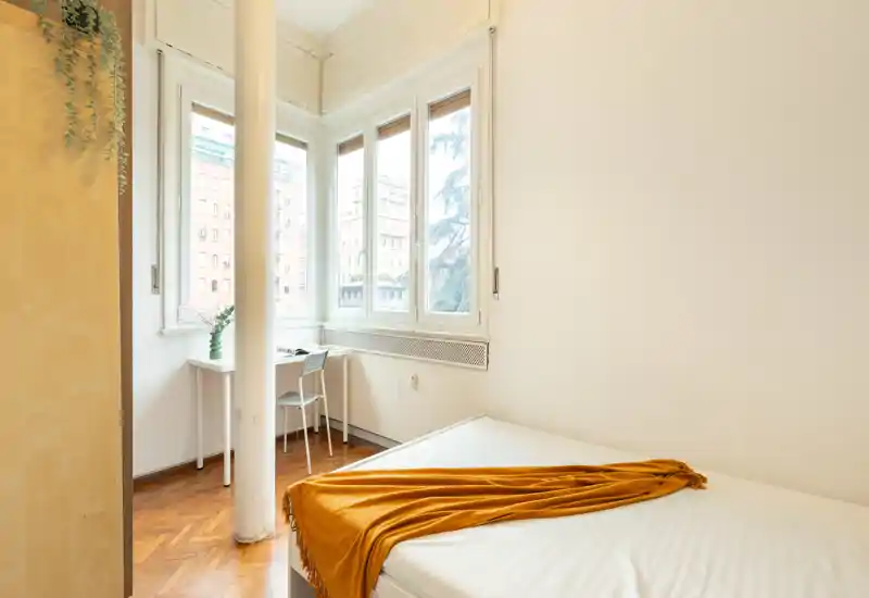 Bright, minimal bedroom with a white mattress, warm ochre throw and a small desk by large windows offering city views. Clean lines and natural light create an airy, inviting space ideal for single occupancy.