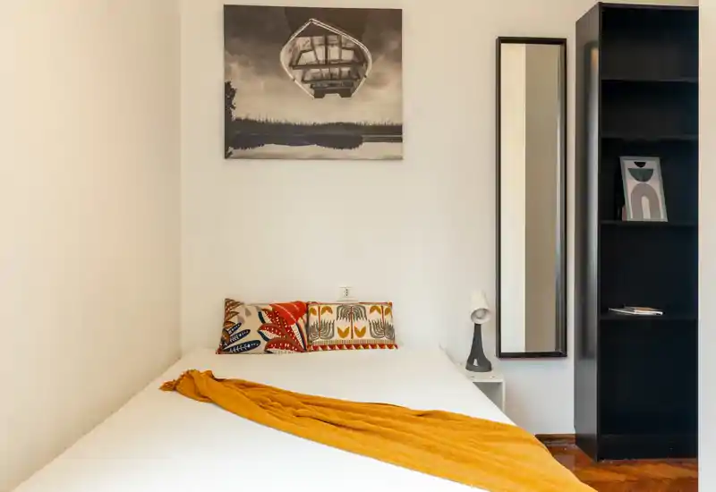 Bright, simply styled bedroom with a double bed, colorful cushions and a warm mustard throw; includes a tall mirror and shelving unit.