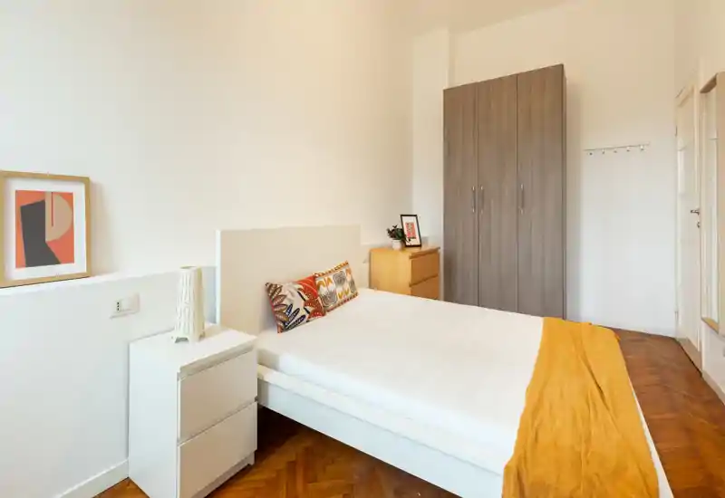 Bright, modern bedroom with a clean white bed, patterned cushions and a warm mustard throw; wardrobe and bedside tables provide practical storage in a minimalist layout.