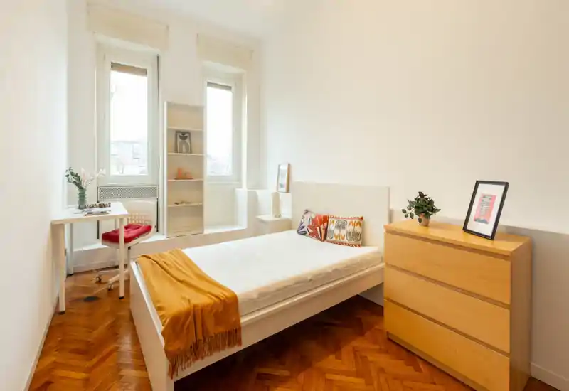 Bright, minimal bedroom with a single bed, wooden parquet floor and a chest of drawers; natural light from two tall windows and a small desk create a cozy, functional space.