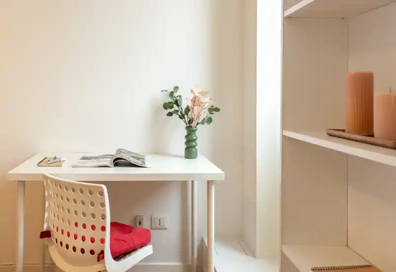 Bright, minimalist workspace/reading nook with a white desk, perforated chair with a red cushion, a green vase with foliage and an open magazine — clean and inviting for short work or reading sessions.