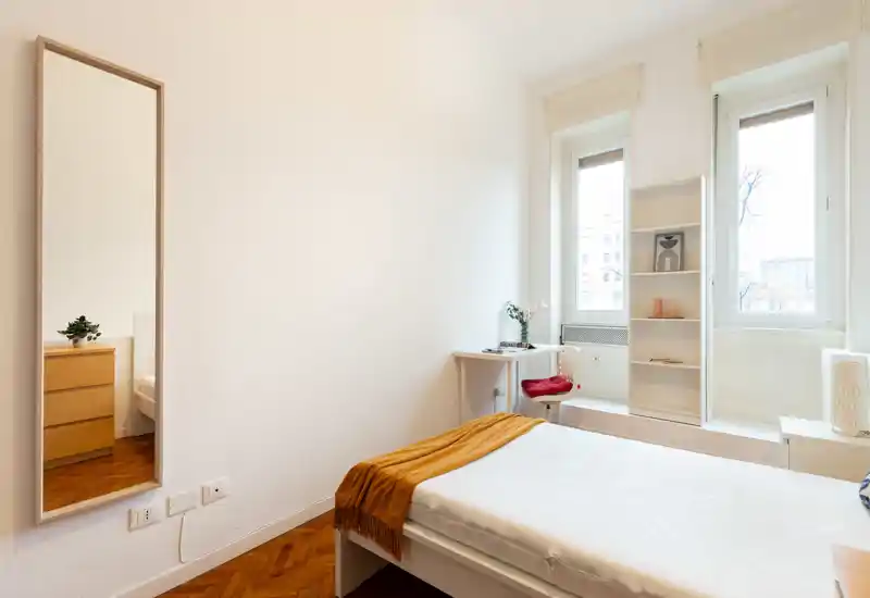 Bright, minimal bedroom with a single bed, warm throw and a small desk by the window. Large windows provide abundant natural light and the room has clean, neutral décor ideal for rental photos.