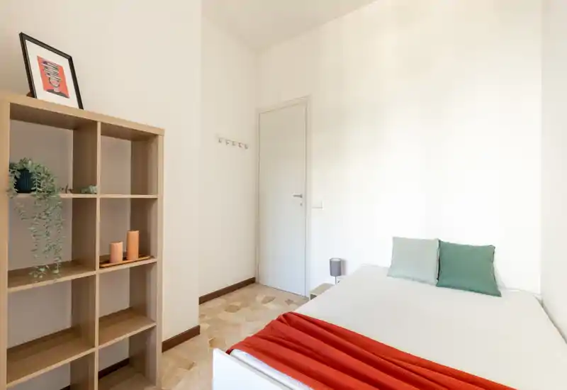 Dormitorio individual luminoso y minimalista con cama bien hecha, cojines de color y manta roja. Una estantería cúbica de madera con plantas y decoración aporta carácter y almacenamiento.