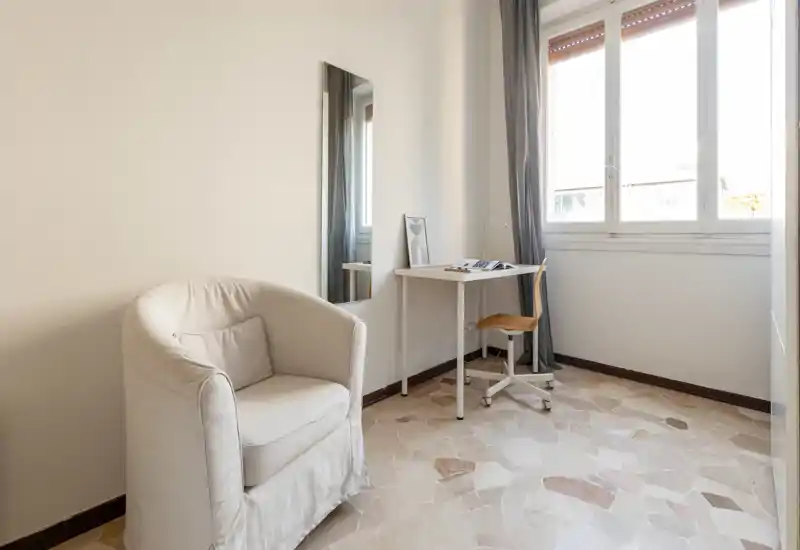 Bright, minimally furnished living area with a comfortable upholstered armchair, a small desk by a large window and neutral decor creating a calm, airy feel.