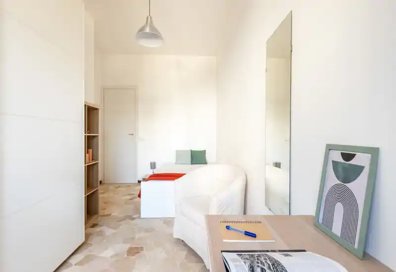 Dormitorio luminoso y minimalista con cama individual, cojines y un pequeño escritorio; tonos neutros y luz natural que crean un ambiente tranquilo y aireado, ideal para una persona.