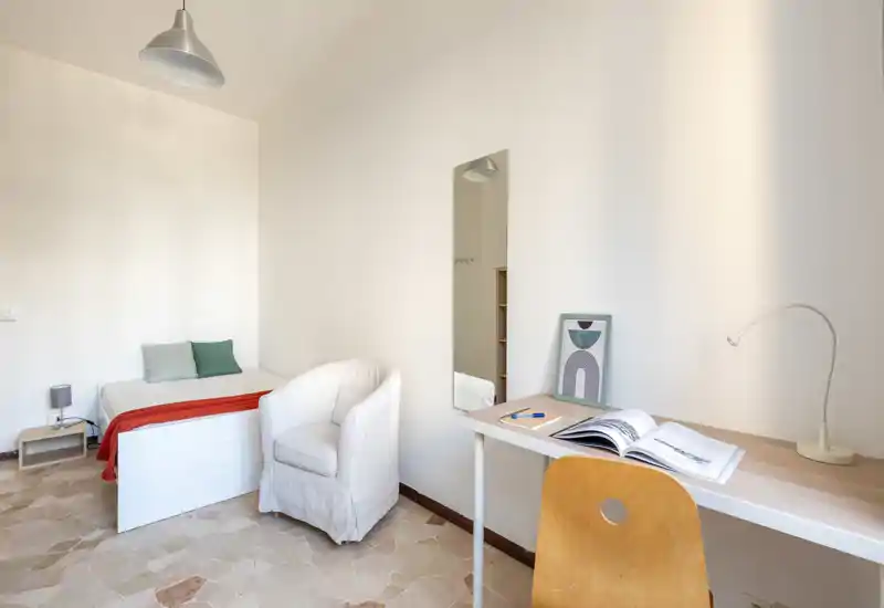 Dormitorio luminoso y minimalista con cama individual blanca, cojines y un sillón acogedor. Escritorio de madera clara con silla y lámpara para un práctico rincón de estudio.