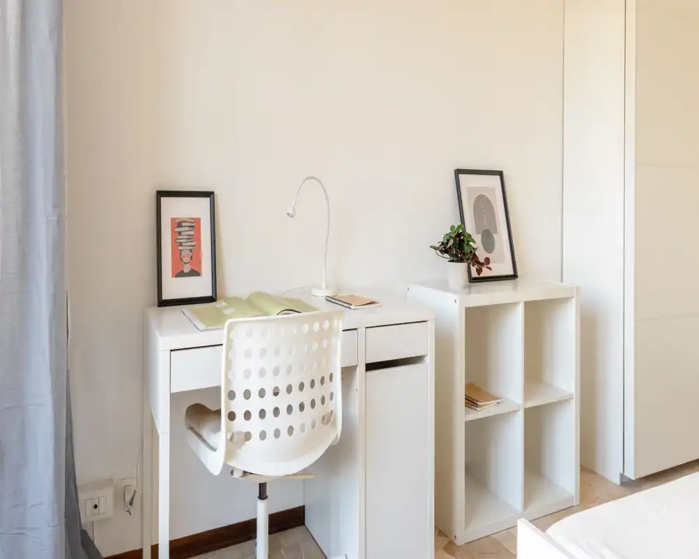 Rincón de dormitorio luminoso y minimalista con escritorio blanco y silla junto a la cama parcialmente visible; cuadros enmarcados y una pequeña estantería crean un ambiente ordenado y sereno.