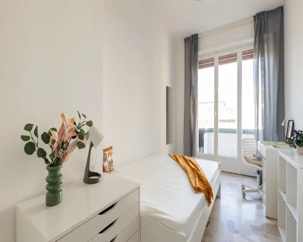 Dormitorio luminoso y minimalista con una cómoda cama individual, textiles claros y balcón con puertas que aportan gran luz natural.