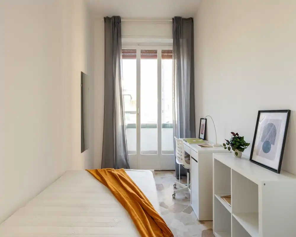Dormitorio luminoso y minimalista con colchón blanco, una manta naranja y un pequeño escritorio junto a la puerta al balcón — ideal como habitación individual compacta y bien iluminada.