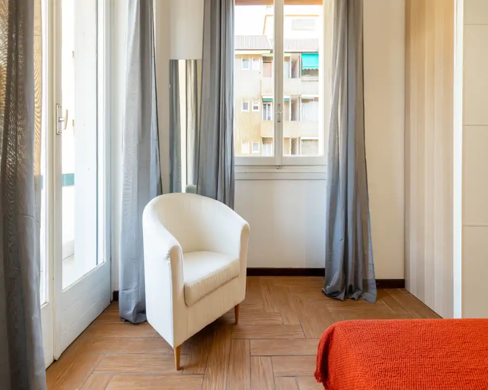 Angolo di camera da letto luminoso e dallo stile semplice con una comoda poltroncina bianca, tende grigie e un accenno di copriletto rosso; la luce naturale dalla finestra e dalla porta del balcone rende l'ambiente accogliente.
