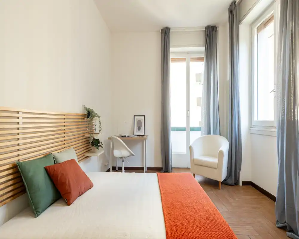 Camera da letto luminosa e moderna con un grande letto, testiera a doghe e un caldo copriletto arancione; luce naturale abbondante, scrivania e poltrona creano un angolo confortevole.