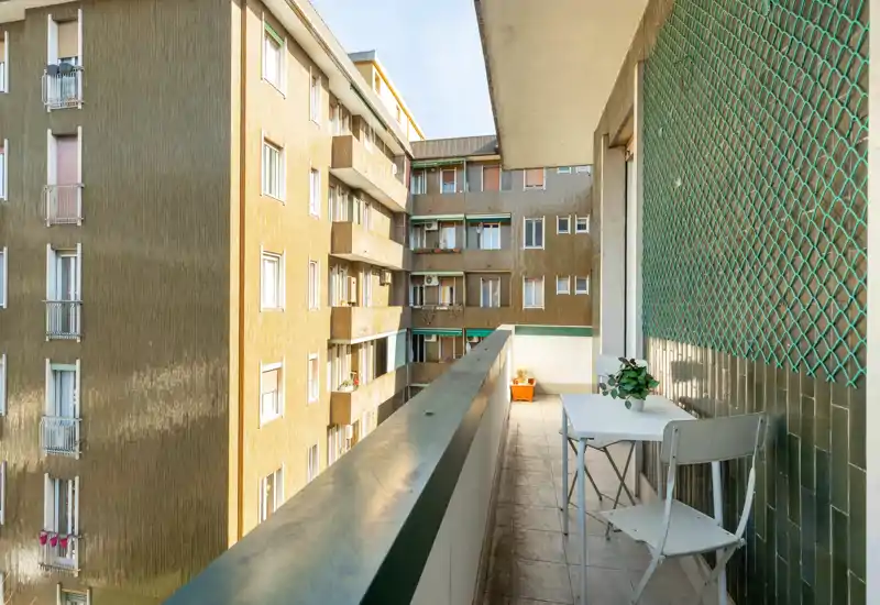Sunny narrow balcony with a small table and chair, decorative plant and view onto neighboring apartment buildings — ideal for morning coffee.