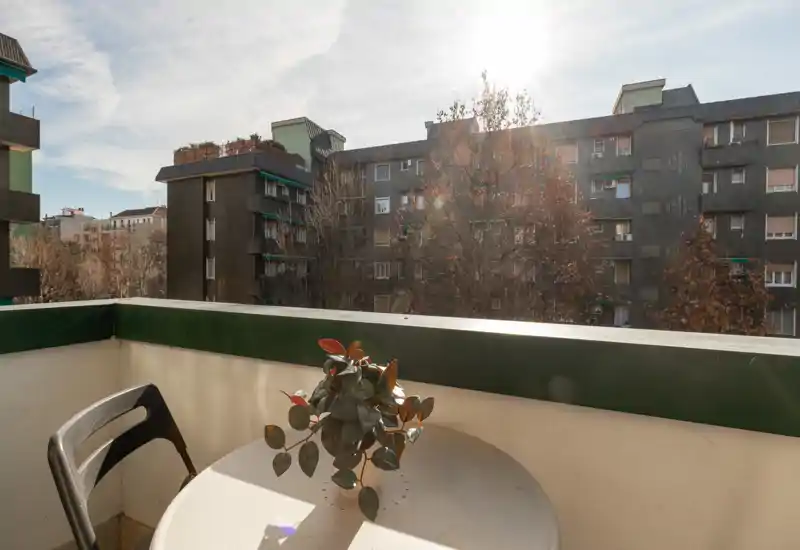 Sunny balcony with a small round table, a potted plant and a chair overlooking neighboring apartment buildings — ideal for morning coffee.