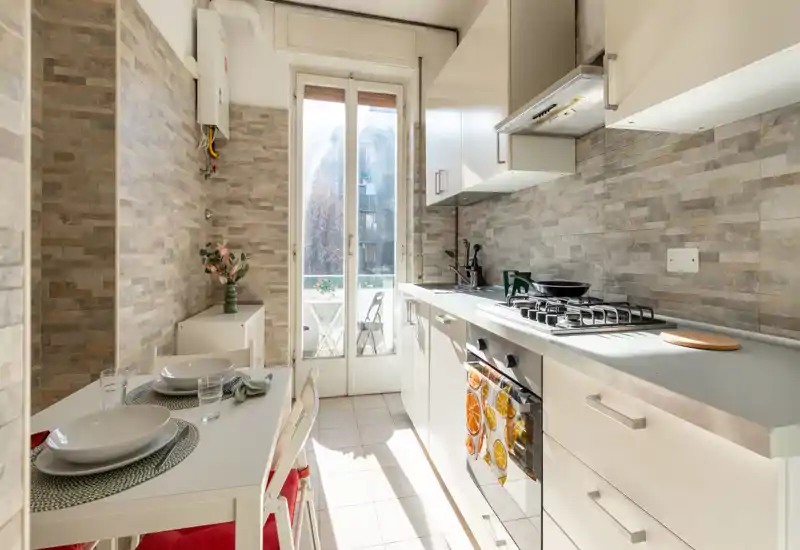 Bright, modern galley kitchen with a gas stove, oven, and a small dining table by the window leading to a balcony. Neutral tile backsplash and white cabinets create a clean, airy feel.