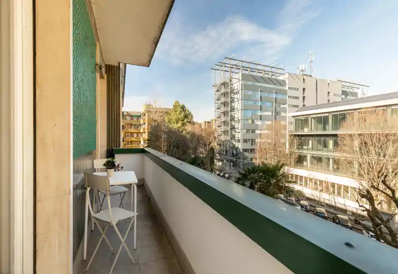 Sunny city balcony with a small bistro table and two chairs, offering street and building views — ideal for morning coffee.