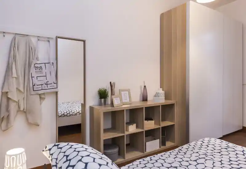 Camera da letto luminosa e accogliente con biancheria a fantasia, mobile contenitore in legno e armadio — ben allestita per annunci.