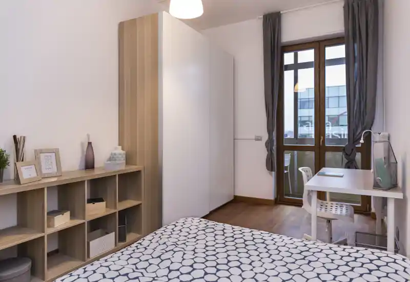 Camera da letto moderna e luminosa con biancheria a motivo, contenitori integrati e una scrivania vicina alla finestra. Pavimento in legno caldo, ampio armadio e luce naturale morbida.
