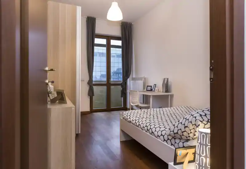Camera singola moderna e luminosa con letto dal motivo geometrico, pavimento in legno e una scrivania vicino alla finestra — ideale per uno studente o un single.
