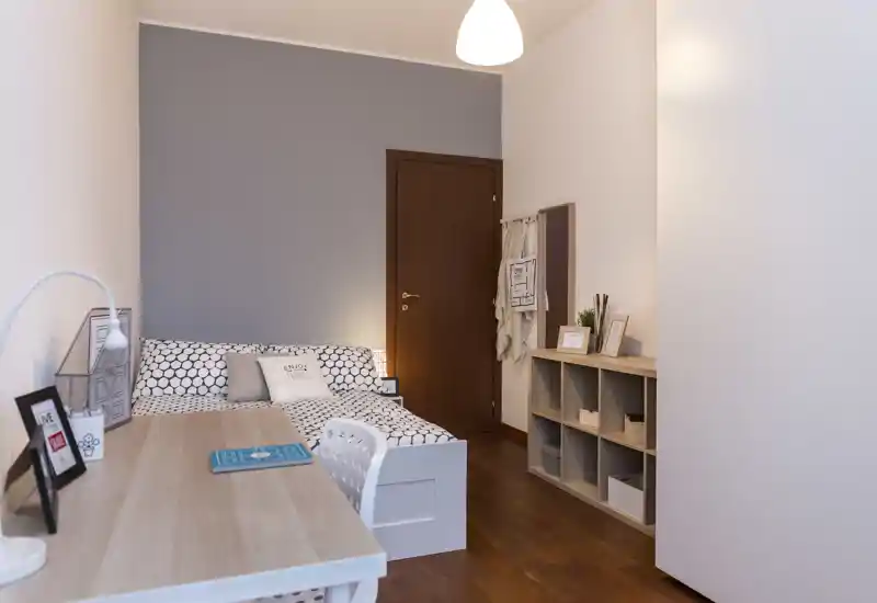 Camera da letto luminosa e moderna con letto dal motivo geometrico, scrivania compatta e scaffalatura — ambiente accogliente e ben illuminato, ideale per una persona o uno studente.