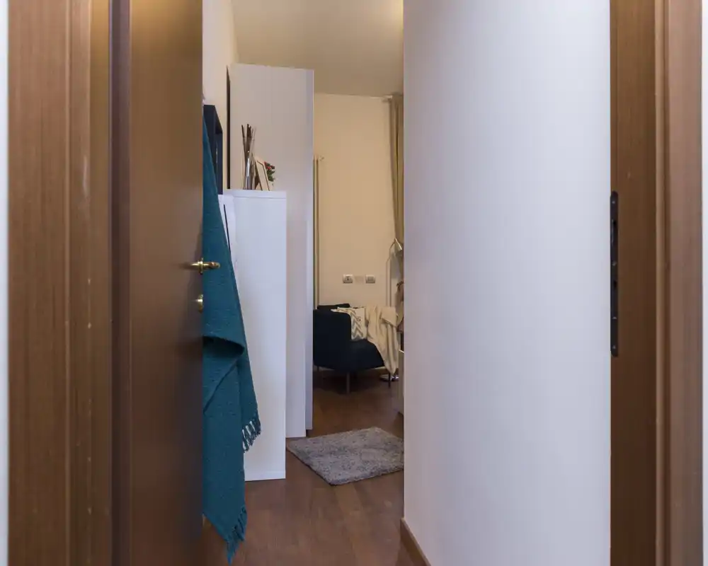 Entrada/pasillo estrecho que conduce a una pequeña zona de estar; suelo de madera, una alfombra y una manta turquesa colgada en la puerta dan una sensación acogedora.