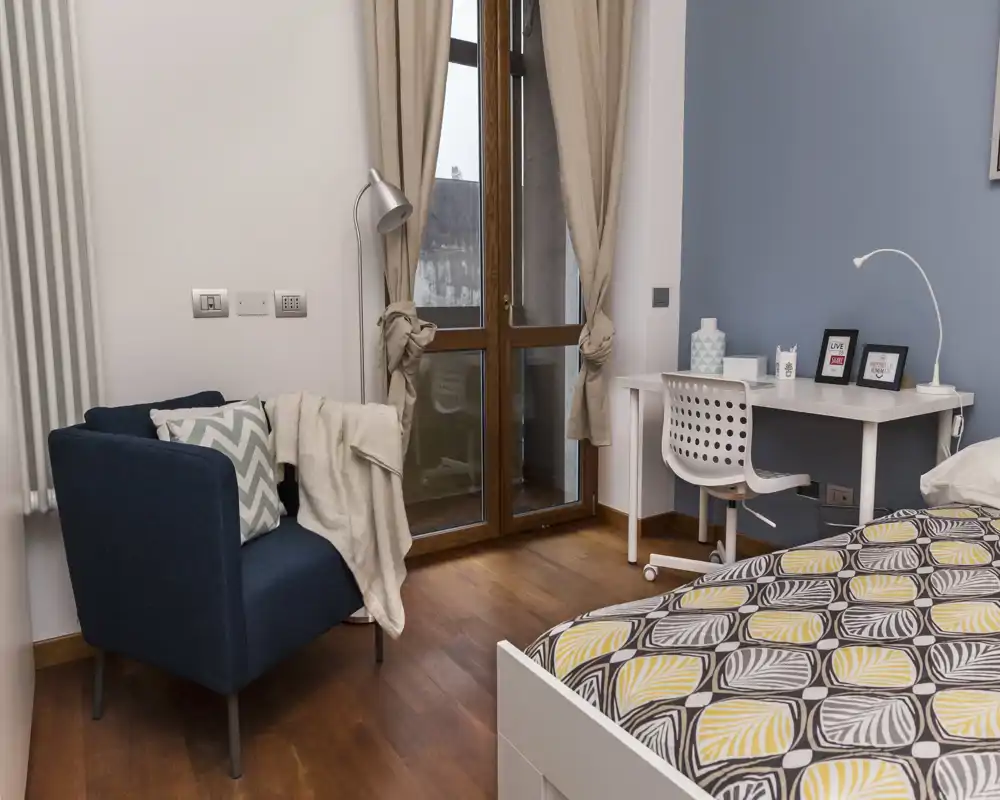 Rincón de dormitorio luminoso y moderno con edredón estampado, un pequeño escritorio blanco y un cómodo sillón azul junto a la ventana. La luz natural y el suelo de madera crean un ambiente acogedor, ideal para alquileres de corta estancia.