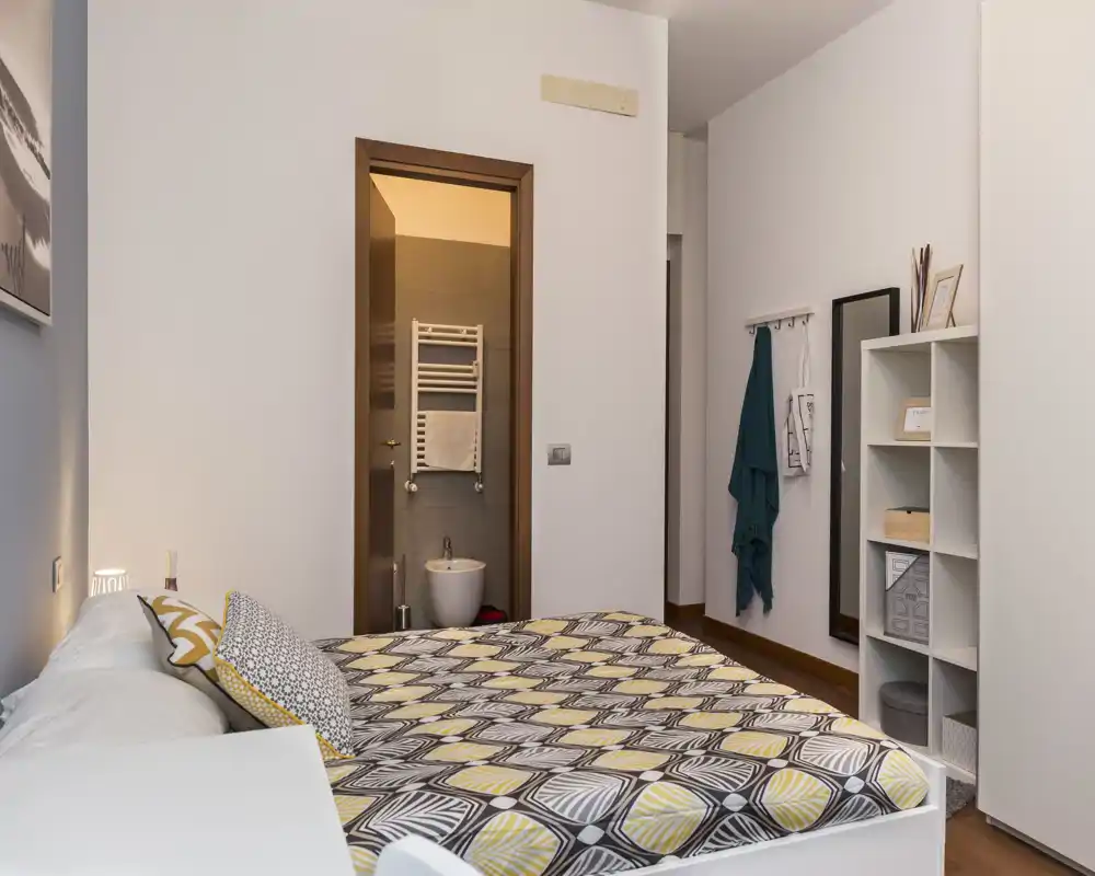 Dormitorio doble luminoso y bien decorado con ropa de cama estampada, lámpara de mesita y baño en suite visible por la puerta; estanterías y percheros aportan funcionalidad.
