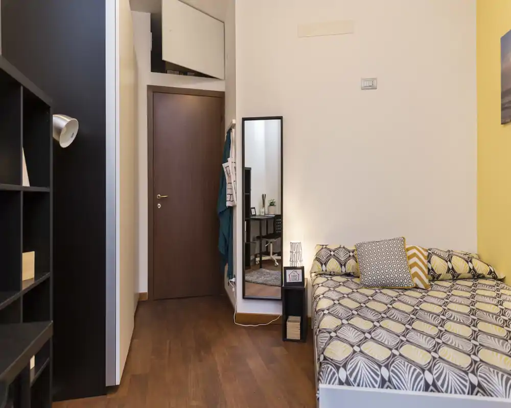 Camera da letto luminosa e curata con biancheria a fantasia, cuscini decorativi e una lampada da comodino; pavimento in legno e un grande specchio completano l'ambiente.