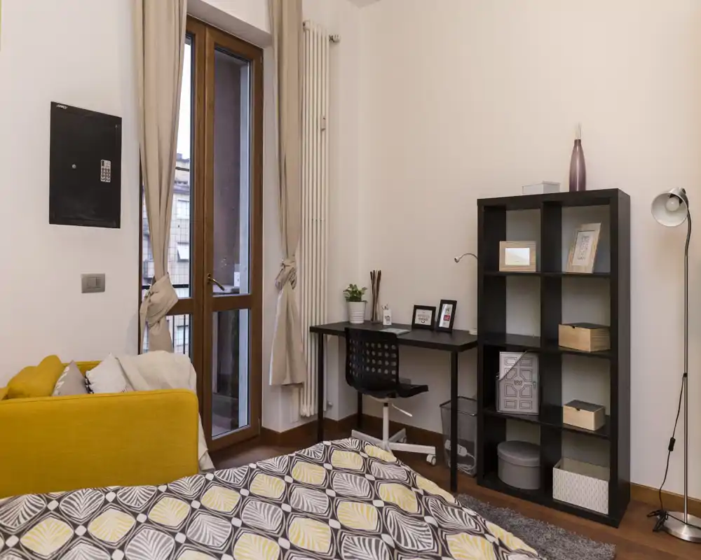 Angolo camera luminoso e accogliente con copriletto decorato, una piccola scrivania e scaffale; pavimento in legno e porta sul balcone che illumina l’ambiente.