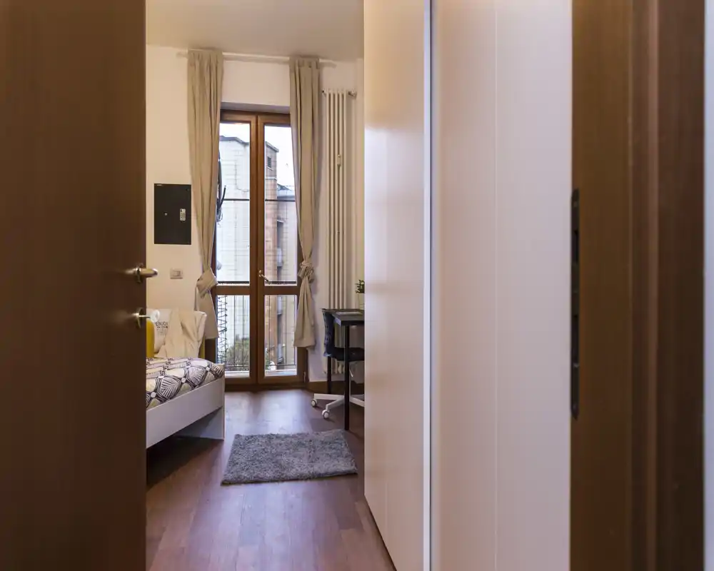 Camera da letto luminosa e semplice con letto singolo, scrivania e porta finestra che lascia entrare luce naturale — ideale per uno studente o una persona sola.