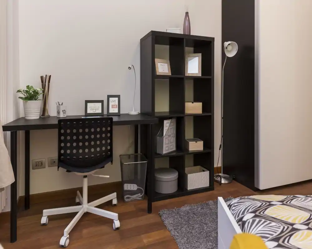 Angolo camera luminoso e moderno con una scrivania, una libreria a cubi e l'angolo del letto con una copertura a motivo. Lo spazio è ordinato e arredato per comfort e produttività.