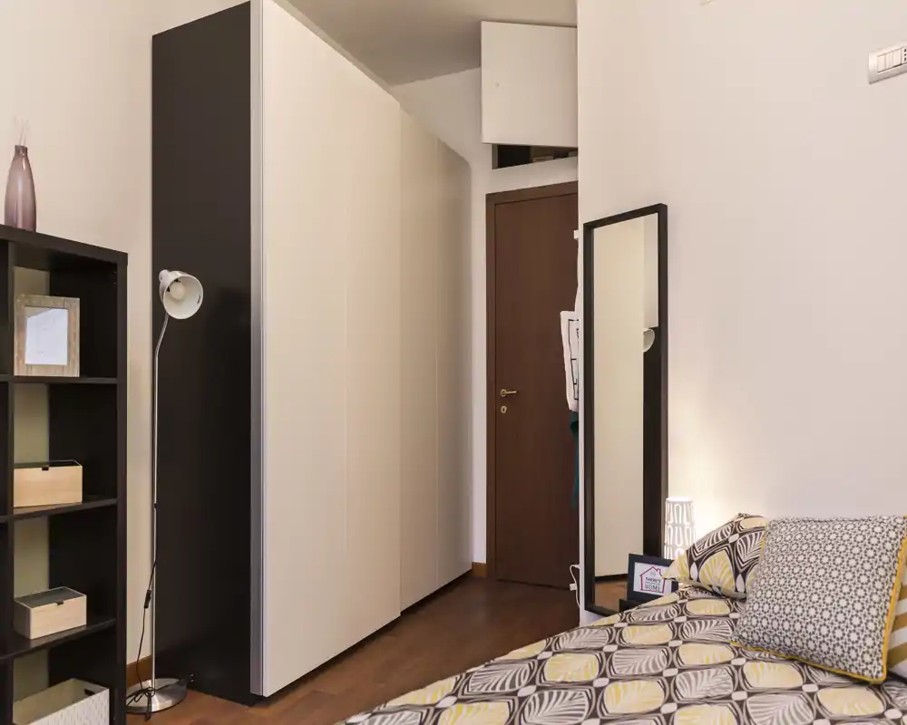 Angolo camera accogliente e ben arredato con biancheria a fantasia, armadio a tutta altezza, lampada da terra e specchio a parete — illuminazione calda e arredamento ordinato, perfetto per un annuncio.
