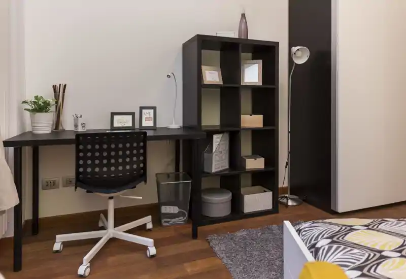 Angolo camera luminoso e ordinato con bordo del letto visibile, scrivania e scaffalatura—ideale per ospiti o uno spazio studio in casa.