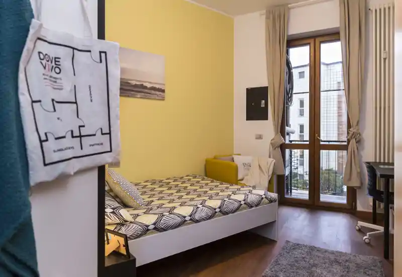 Camera singola luminosa e accogliente con biancheria a motivi, piccola poltrona e ampia finestra che lascia entrare luce naturale — ideale per studente o single.