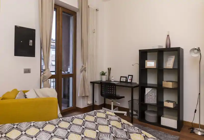 Camera da letto luminosa e accogliente con piumone a motivi, una piccola scrivania e una libreria; la luce naturale dalla porta del balcone crea un’atmosfera calda.
