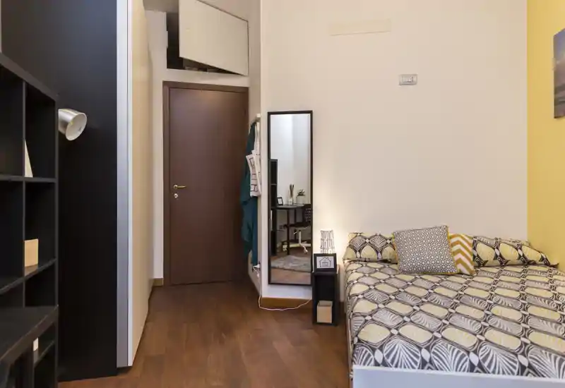 Camera da letto luminosa e accogliente con biancheria a fantasia, cuscini decorativi e specchio a figura intera; pavimento in legno e illuminazione calda.