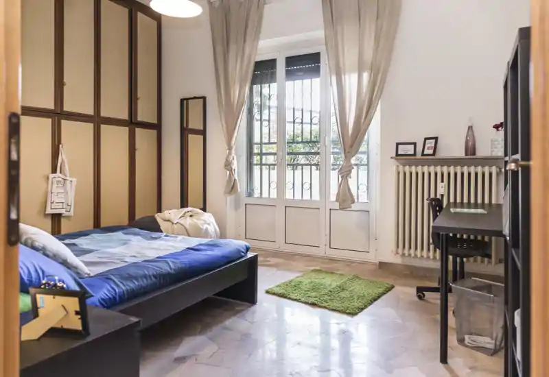 Camera da letto luminosa e ben arredata con letto matrimoniale, ampia finestra che illumina l'ambiente, scrivania e armadio — ideale per annunci in affitto che evidenziano comfort e area studio.