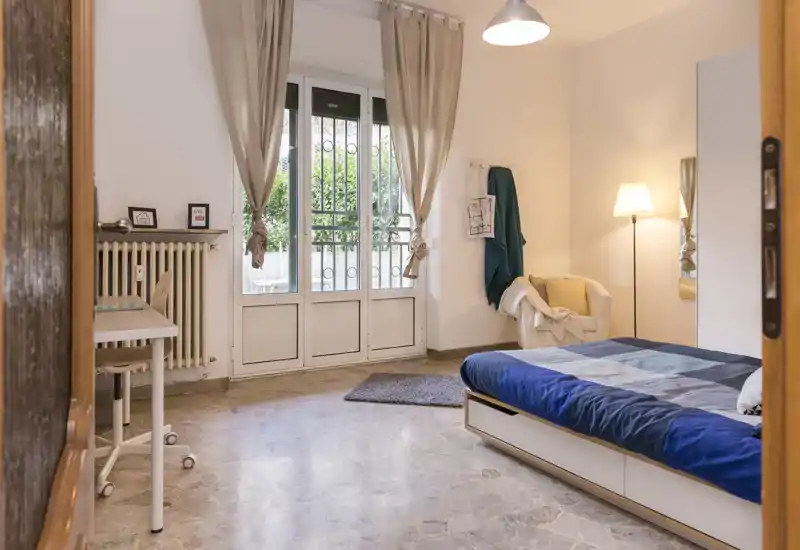 Camera da letto luminosa e arredata con letto matrimoniale, ampia finestra con tende, poltrona per la lettura e scrivania — ideale per soggiorni comodi.