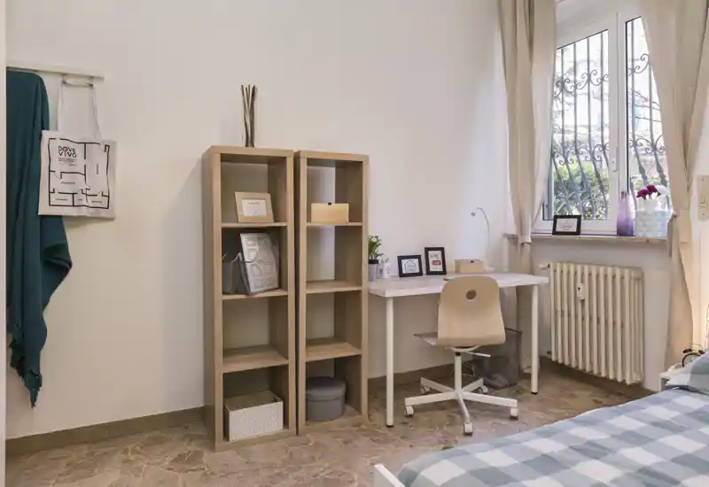 Angolo camera luminoso e accogliente con una scrivania vicino alla finestra, scaffalatura aperta e parte del letto visibile con copriletto a quadri.