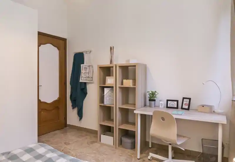 Angolo della camera ordinato e ben illuminato con letto visibile, scaffali in legno e una scrivania ordinata ideale per il lavoro da remoto.