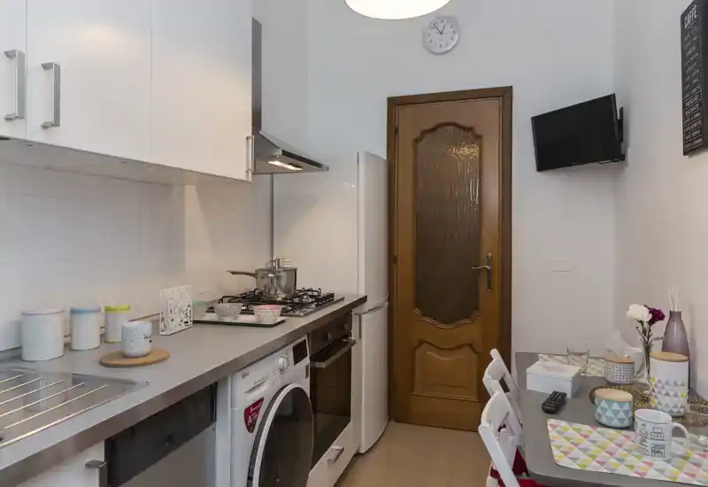 Cucina compatta e ben attrezzata con piano cottura a gas, forno, frigorifero e piano d'appoggio con sedute. Luminosa, ordinata e decorata con tazze colorate e tovagliette per un'atmosfera accogliente.