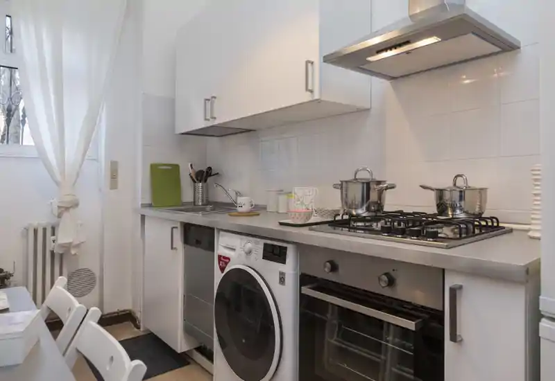 Cucina moderna compatta con piano cottura a gas in acciaio, forno incassato e lavatrice, mobili bianchi e paraschizzi piastrellato. La luce naturale dalla finestra illumina l'area funzionale e pulita.