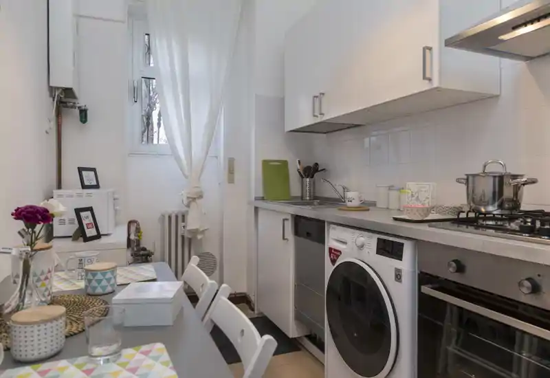 Cucina compatta e ben curata con elettrodomestici integrati, piano cottura a gas e lavatrice/asciugatrice sotto il piano. Un piccolo tavolo è apparecchiato con tovagliette colorate e un vaso di fiori, creando un ambiente accogliente e funzionale.