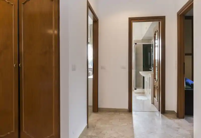 Ingresso luminoso con pavimento in marmo, porte e armadiatura in legno, che conduce alle stanze adiacenti incluso un bagno visibile. Arredamento pulito e neutro, perfetto per un annuncio immobiliare.