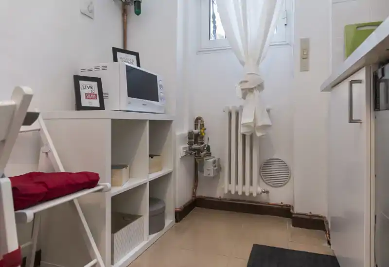 Angolo cucina compatto con scaffalatura bianca, microonde e piccolo piano di lavoro, illuminato da una finestra con tenda; ordinato e funzionale per preparazioni veloci.