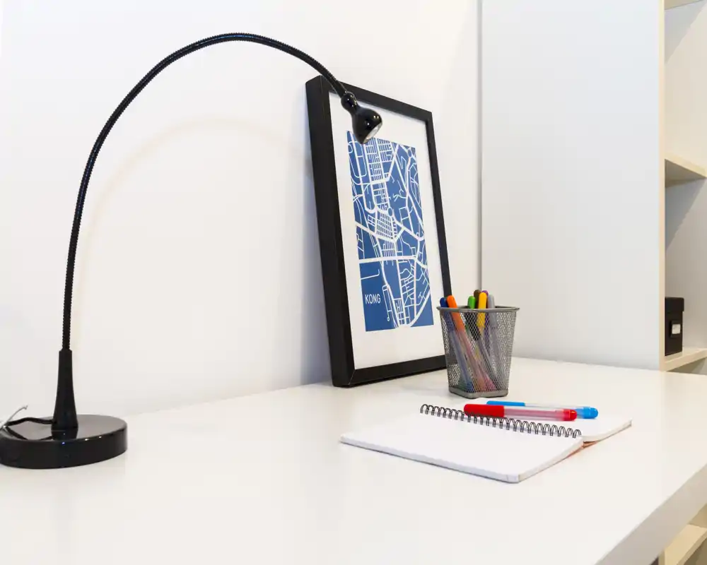 Angolo scrivania minimal e luminoso con una lampada nera regolabile, stampa incorniciata e cancelleria su una superficie bianca — perfetto per un piccolo spazio di lavoro.